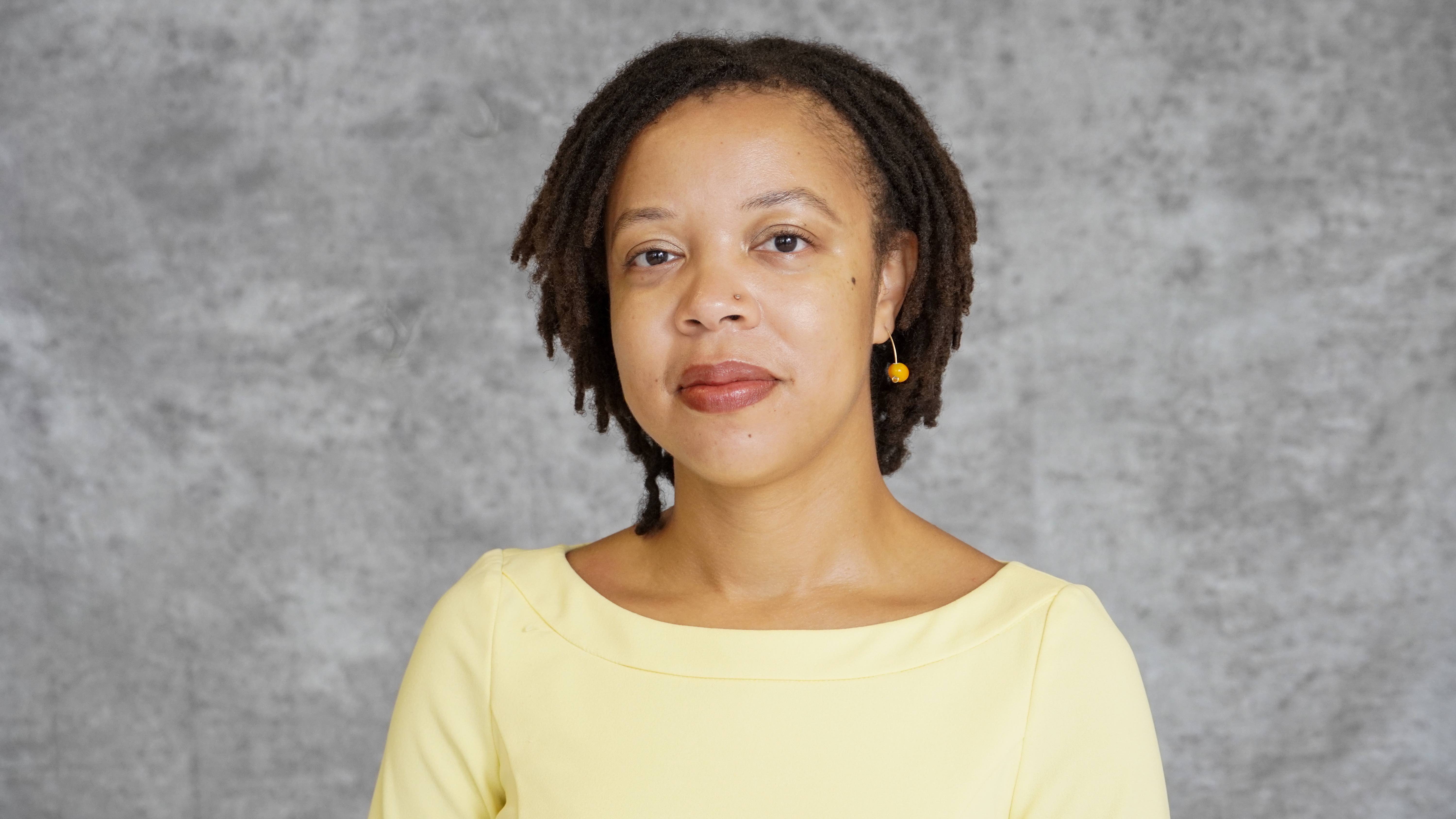 Headshot image of Dr. Amelia Simone Herbert. She has short locs and a yellow dress and looks directly into the camera.